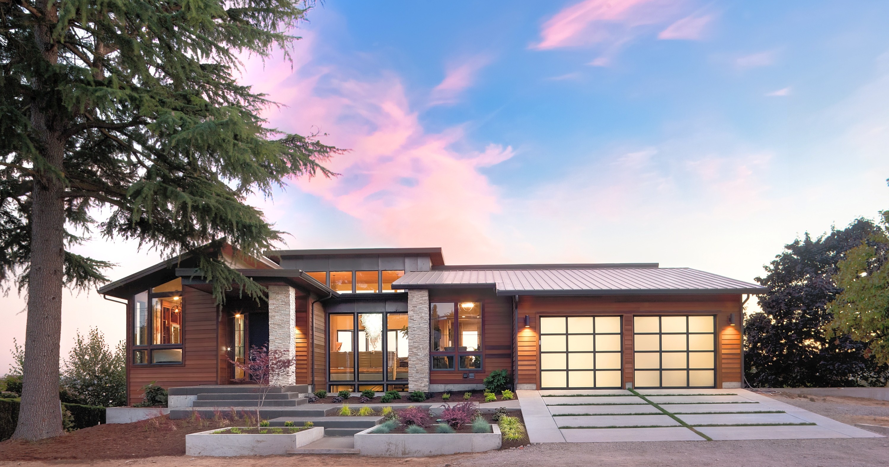 A modern Pacific-Northwest custom home at dusk, its interior warmly lit among tall evergreens — built by Custom Royal Homes