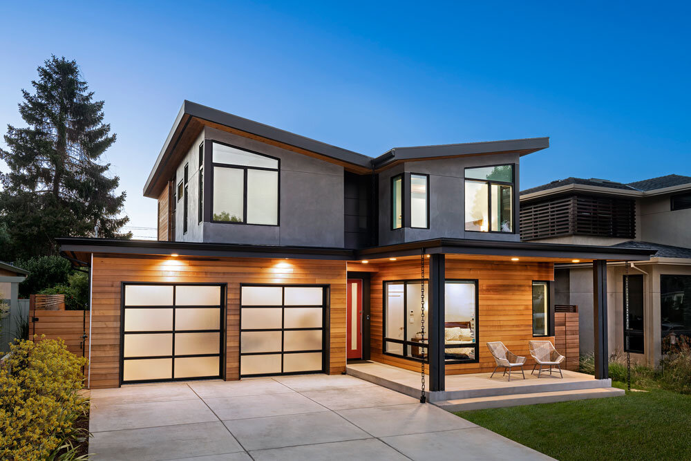 A modern two-story custom home at dusk with warm lighting and glass garage doors